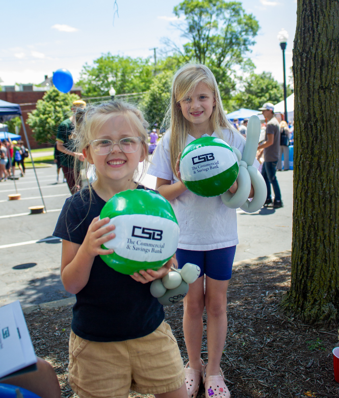 Two smiling kids holding CSB beach balls and balloon animals at community event